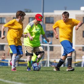 Two athletes in yellow t-shirts and blue shorts playing soccer against an athlete in a green t-shirt and tan shorts.