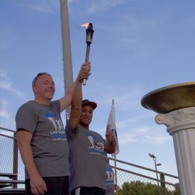 Two males wearing grey t-shirts standing outside holding a torch together.