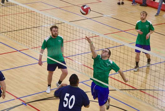 Volleyball players compete at the net during an indoor match, with one player reaching to return the ball