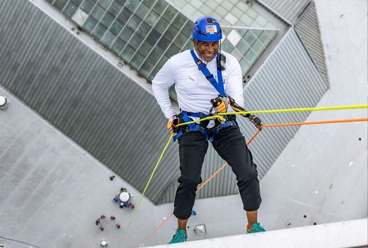 Woman in climbing gear and a helmet climbing down a building holding two thumbs up.