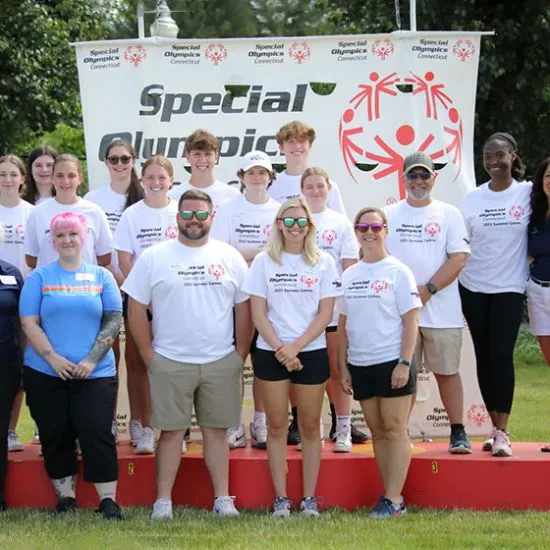 Men and women standing outside wearing white t-shirts in front of a Special Olympics banner.
