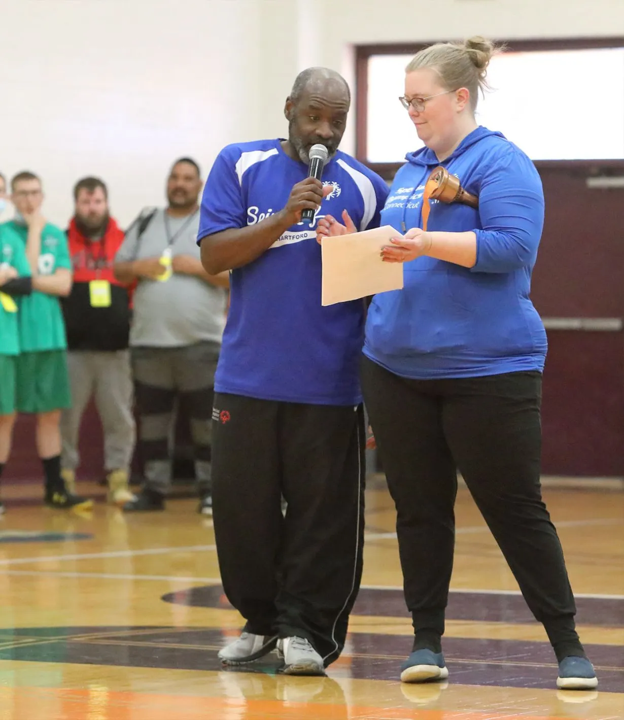 Male and female standing in the middle of a gymnasium wearing blue tops and speaking into a microphone.