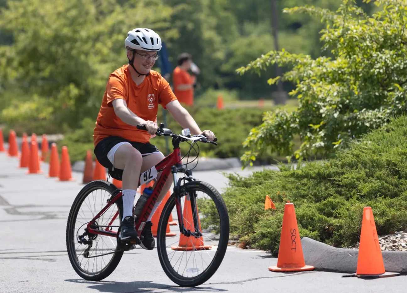 Athlete on a bike wearing an orange shirt near green trees.