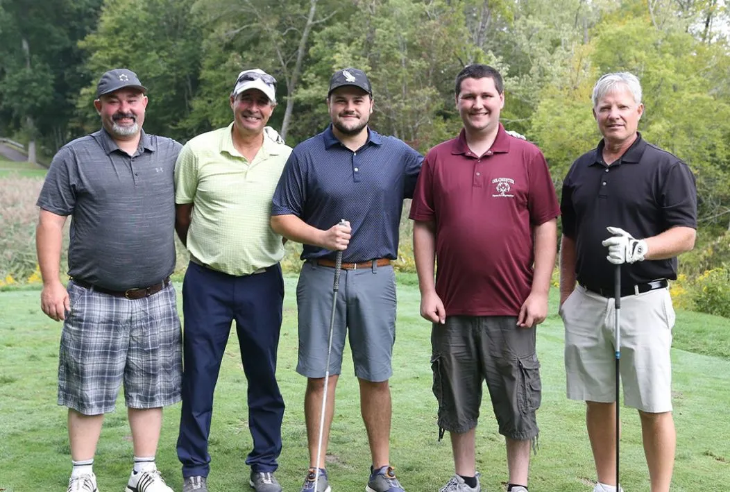 Five men standing together on a golf course holding golf clubs and smiling.
