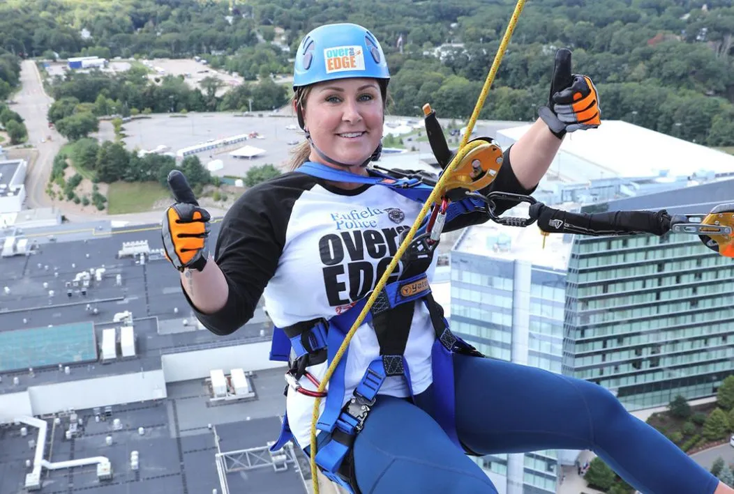 Woman in climbing gear and a helmet climbing down a building holding two thumbs up.