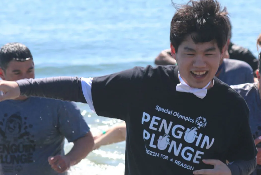 Young man running in the water with others wearing a bowtie and a black shirt with the Penguin Plunge logo.