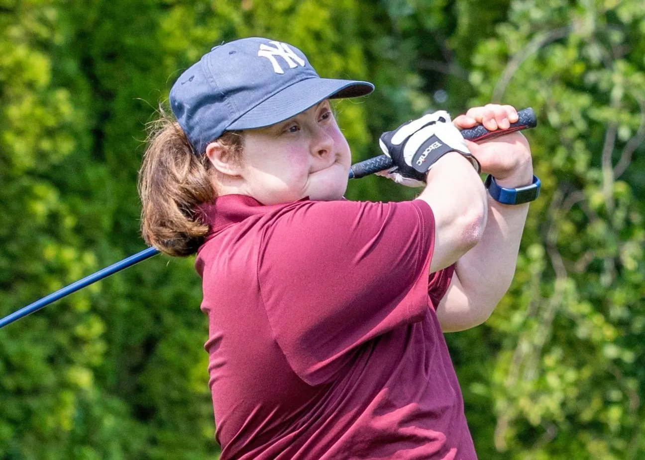 Female athlete in maroon golf shirt and New York Yankees hat holding her golf stroke watching the ball land.