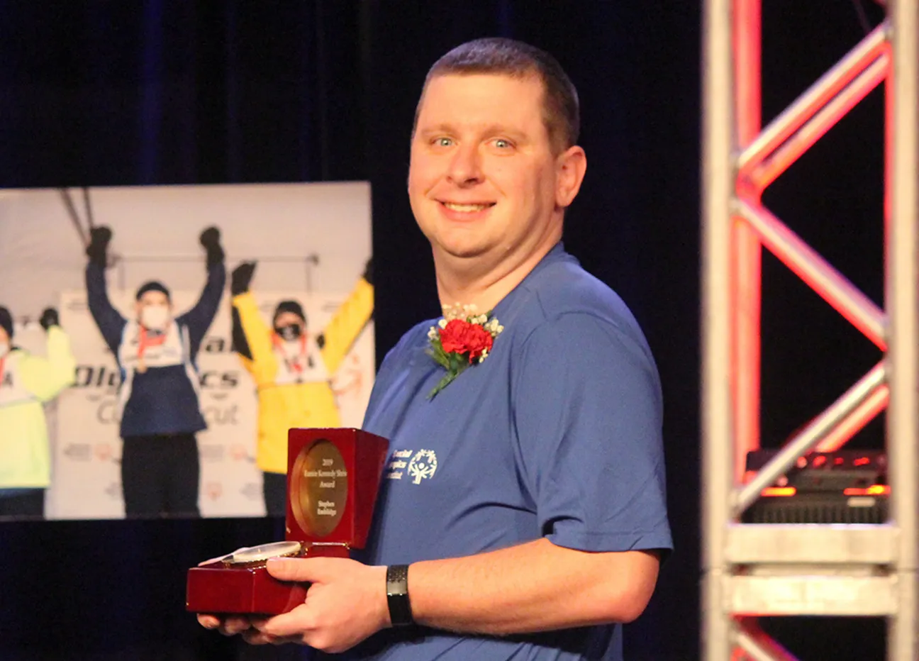 Male wearing a blue t-shirt smiling and holding an award.