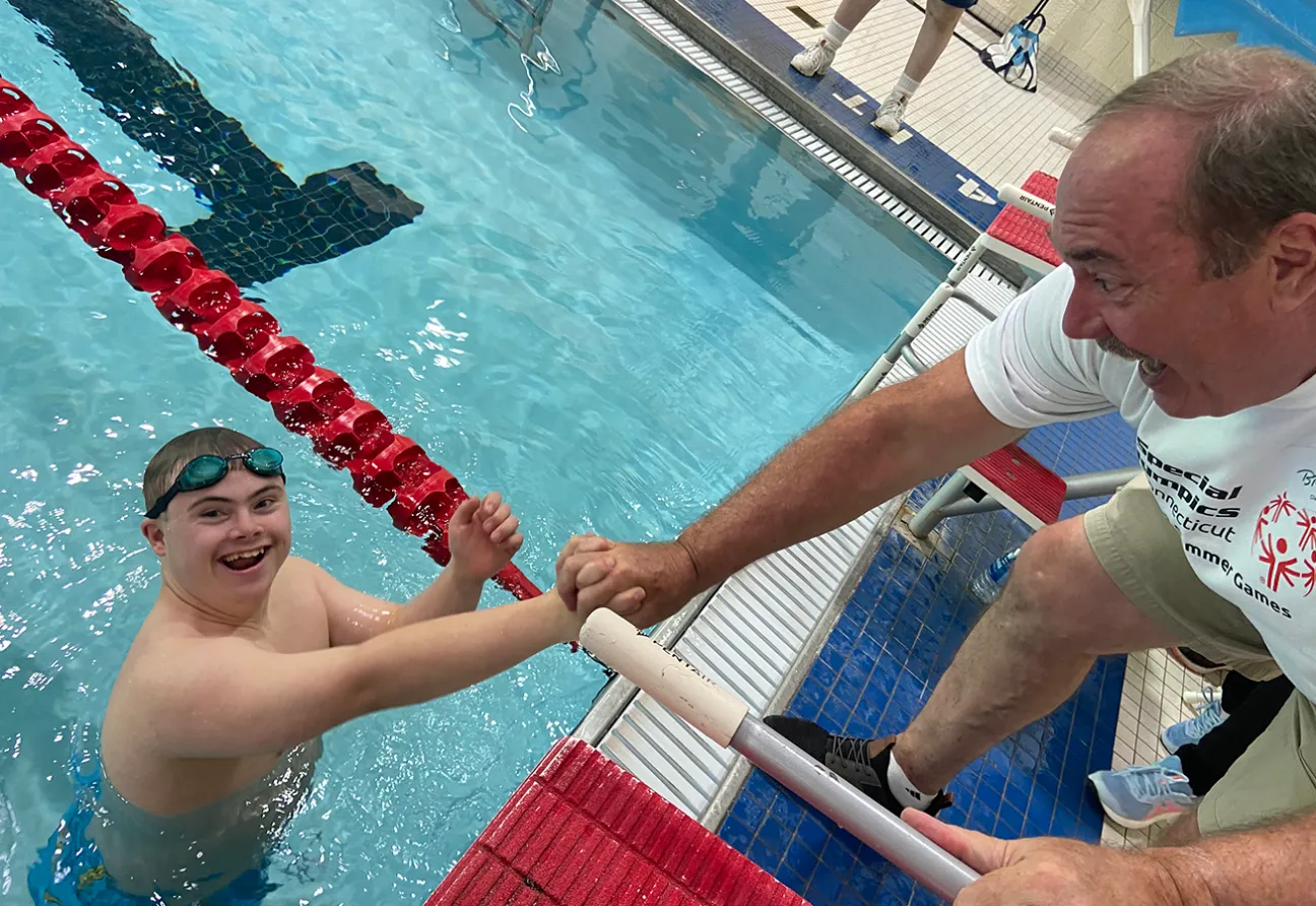 Athlete standing in a pool with goggles on his head smiling and holding his coach's hand.