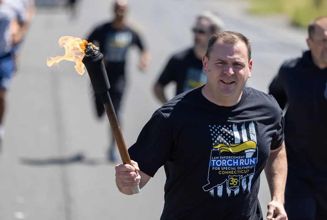 Man carrying torch wearing a black torch run shirt jogging with others.
