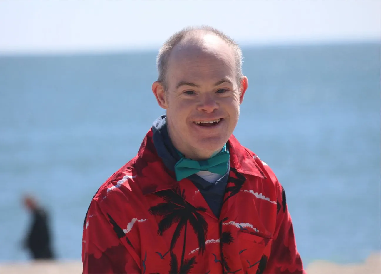 Male smiling by a body of water wearing a teal bowtie and red tropical shirt.