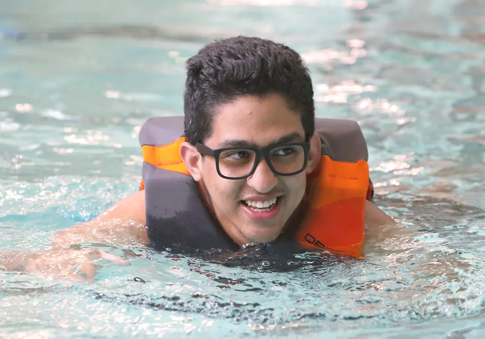 Young boy wearing glasses swimming in pool with life vest.