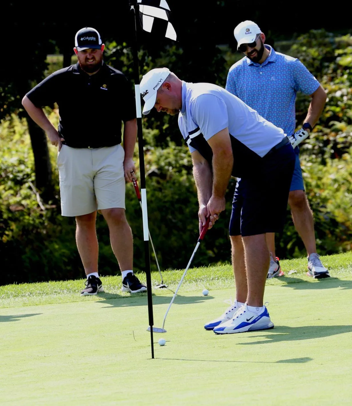 One man playing golf on a golf course while two other men look on from the side.